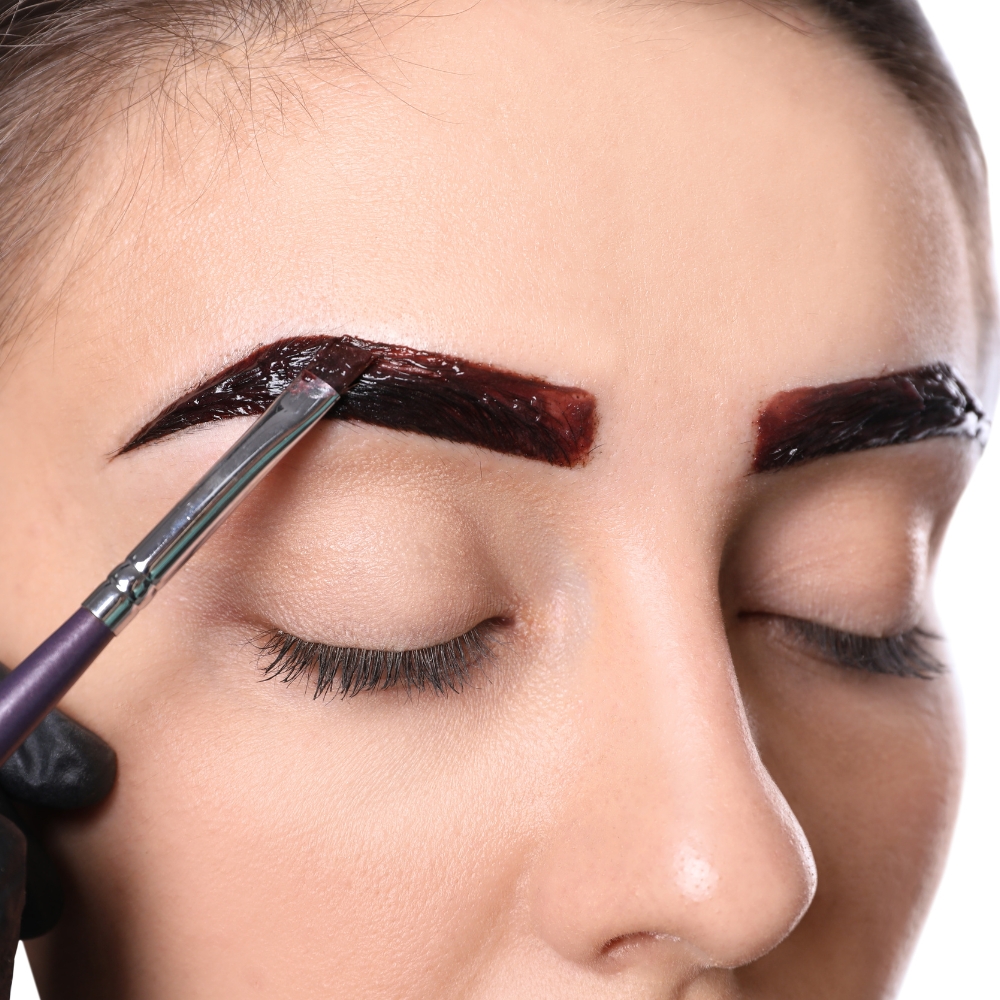 The image captures the process of a brow wax and tint, where a dark tinting solution is applied to the eyebrows, and excess hair is shaped and removed for a clean, defined look. This treatment enhances the natural brow shape and color, offering a polished finish.