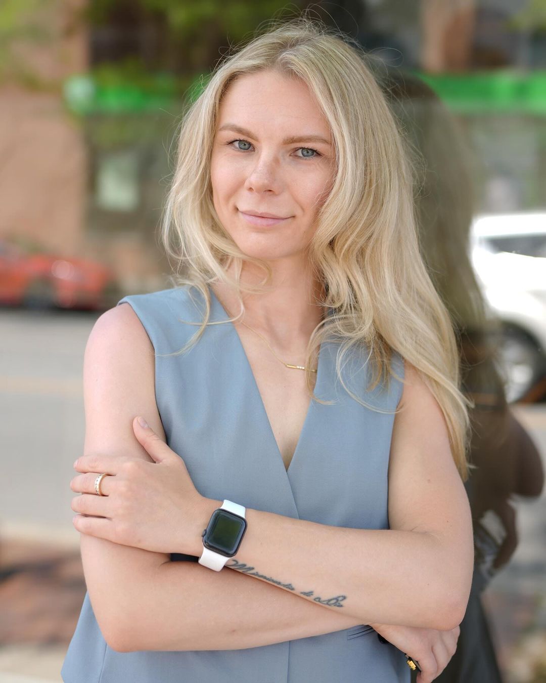 The image features the owner of a business, presenting a confident and relaxed stance with her arms crossed. She is standing outdoors, with a street and greenery in the background, which creates a pleasant urban setting. The owner's gentle smile and casual pose, combined with her professional attire, convey both approachability and professionalism.